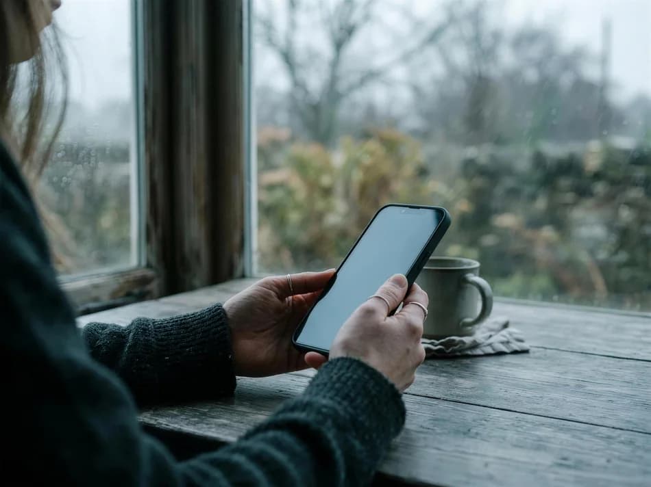 Eine Person hält an einem grauen Tag ein Smartphone über einen Tisch am Fenster