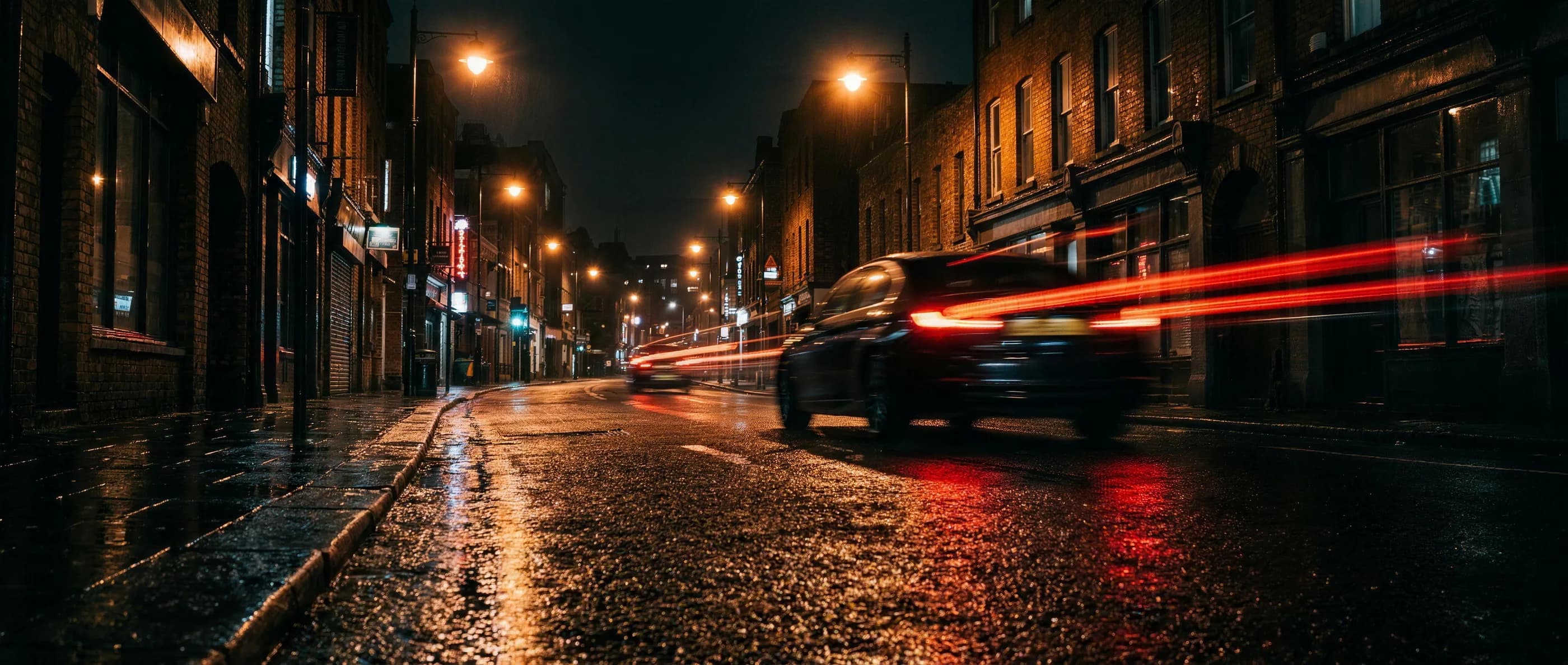 Nasse Stadtstraße bei Nacht, ein Auto fährt mit langen roten Rücklichtspuren vorbei